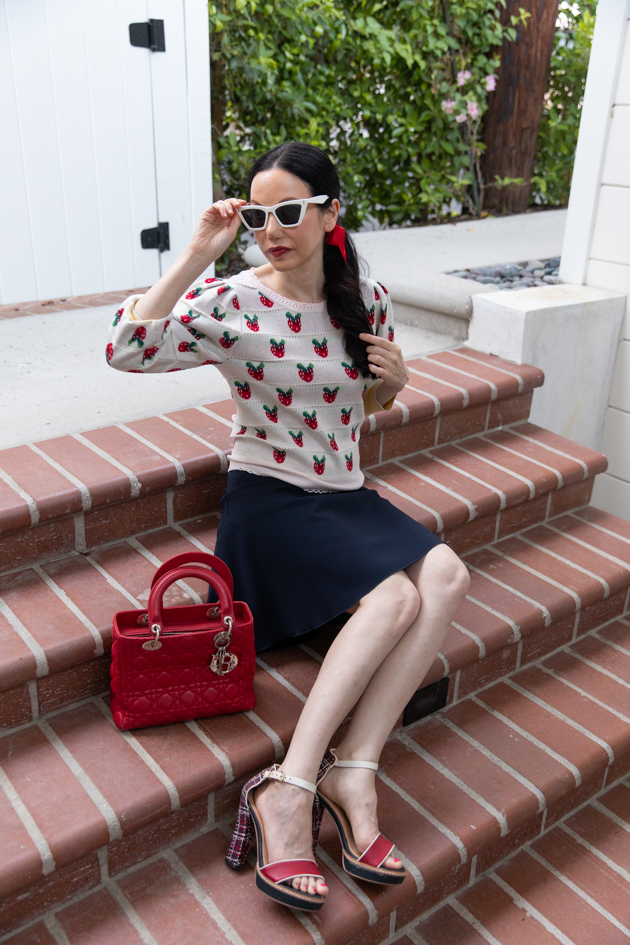 Fall Transitional Look, Waiting for Election Results, Red White and Blue Outfit, LoveShackFancy Strawberry Sweater, Tommy Hilfiger Collection Skirt, Italic Cateye Sunglasses, Lady Dior Bag, Classic and Feminine Style, What to Wear for Fall, Fall in Los Angeles, Preppy Style Los Angeles Lifestyle Blogger, Pretty Little Shoppers Blog #FallTransitionalLook #PreppyStyle | Fall Clothing by popular L.A. fashion blog, Pretty Little Shoppers: image of a woman sitting on some brick steps and wearing a strawberry Net-A-Porter sweater, Dior purse, Tommy Hilfiger skirt, plaid block heel sandals, and Italic sunglasses.