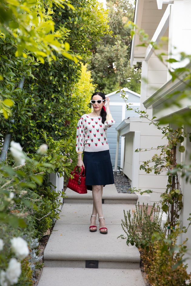 Fall Transitional Look, Waiting for Election Results, Red White and Blue Outfit, LoveShackFancy Strawberry Sweater, Tommy Hilfiger Collection Skirt, Italic Cateye Sunglasses, Lady Dior Bag, Classic and Feminine Style, What to Wear for Fall, Fall in Los Angeles, Preppy Style Los Angeles Lifestyle Blogger, Pretty Little Shoppers Blog #FallTransitionalLook #PreppyStyle |Fall Clothing by popular L.A. fashion blog, Pretty Little Shoppers: image of a woman wearing a strawberry Net-A-Porter sweater, Dior purse, Tommy Hilfiger skirt, plaid block heel sandals, and Italic sunglasses.