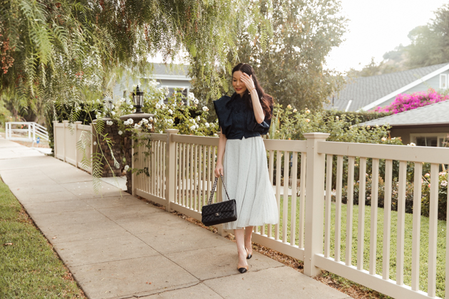 Woman wearing blue statement top and pleated skirt walking on sidewalk |Life Update by popular LA lifestyle blog, Pretty Little Shoppers: image of a woman walking on a sidewalk and wearing a By Abigal Love blue Statement blouse, grey pleated skirt and carrying a Black quilted Chanel bag 