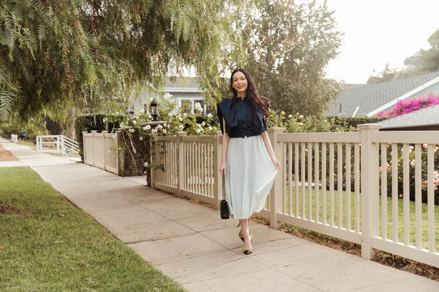 By Abigail Love blue statement blouse with white pleated skirt |Life Update by popular LA lifestyle blog, Pretty Little Shoppers: image of a woman standing next to a white picket fence and wearing a By Abigal Love blue Statement blouse, grey pleated skirt and carrying a Black quilted Chanel bag.