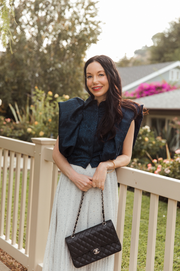 Woman wearing By Abigail Love statement blouse and holding black Chanel bag leaning on white picket fence |Life Update by popular LA lifestyle blog, Pretty Little Shoppers: image of a woman standing next to a white picket fence and wearing a By Abigal Love blue Statement blouse, grey pleated skirt and carrying a Black quilted Chanel bag.