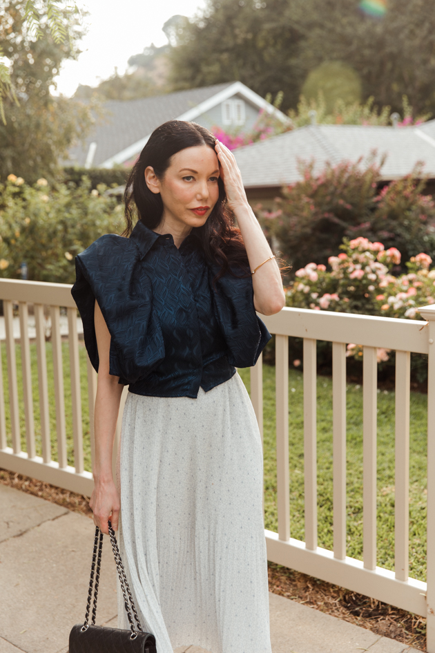 Woman wearing blue statement top on a Spring afternoon |Life Update by popular LA lifestyle blog, Pretty Little Shoppers: image of a woman standing next to a white picket fence and wearing a By Abigal Love blue Statement blouse, grey pleated skirt and carrying a Black quilted Chanel bag.