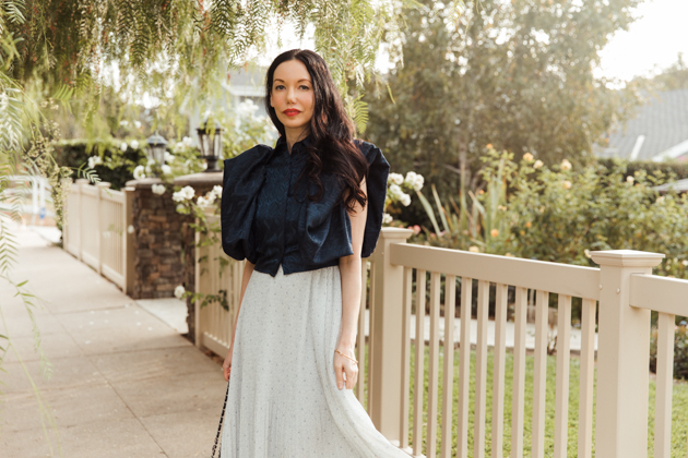 Woman wearing blue top and white pleated skirt stands in front of white picket fence. |Life Update by popular LA lifestyle blog, Pretty Little Shoppers: image of a woman standing next to a white picket fence and wearing a By Abigal Love blue Statement blouse, grey pleated skirt and carrying a Black quilted Chanel bag 