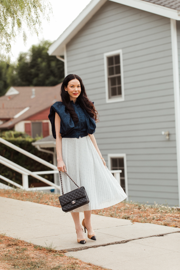 White pleated dress styled with blue statement top |Life Update by popular LA lifestyle blog, Pretty Little Shoppers: image of a woman walking on a sidewalk and wearing a By Abigal Love blue Statement blouse, grey pleated skirt and carrying a Black quilted Chanel bag 