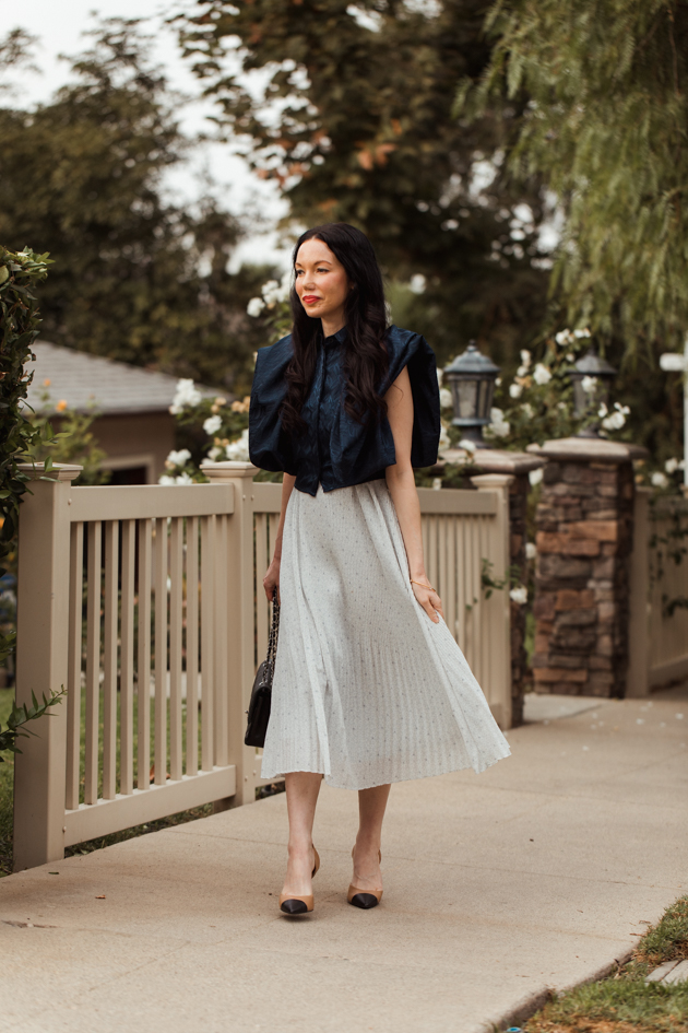 Blue Statement Blouse styled with a pleated skirt for Spring |Life Update by popular LA lifestyle blog, Pretty Little Shoppers: image of a woman standing next to a white picket fence and wearing a By Abigal Love blue Statement blouse, grey pleated skirt and carrying a Black quilted Chanel bag.