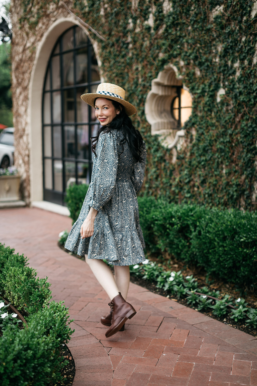 Cottagecore Style, Romantic Dress, Boater Hat |  Romantic Dress by popular Dallas fashion blog, Pretty Little Shoppers: image of a woman walking on a brick pathway next to a building covered in ivy and wearing a Doen Leda fawn dress, Lack of Color straw boater hat, and Suzanne booties.