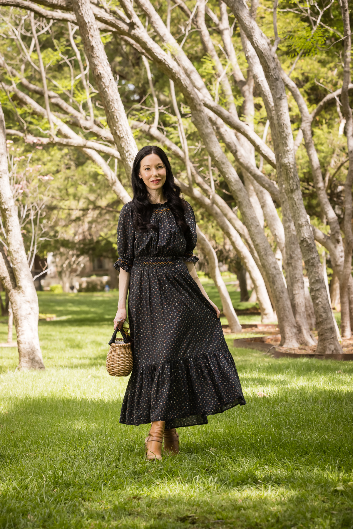 Doen Prairie Dress, Pixie Market Straw Satchel | Prairie Dress by popular Dallas fashion blog, Pretty Little Shoppers: image of a woman walking outside on some grass under some trees and wearing a black Down prairie dress, tan boots, and holding a woven handbag. 