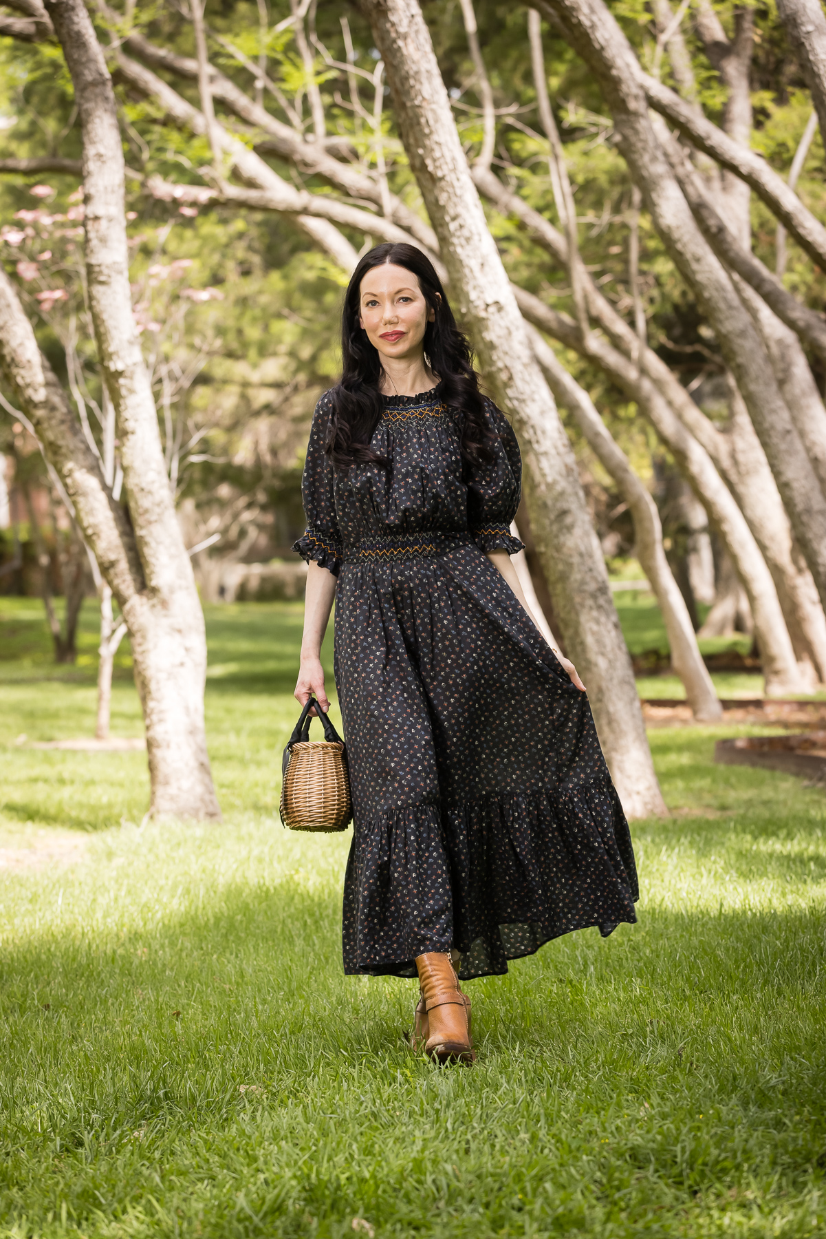 Black Floral Prairie Dress, Straw bag, distressed ankle boots | Prairie Dress by popular Dallas fashion blog, Pretty Little Shoppers: image of a woman walking outside on some grass under some trees and wearing a black Down prairie dress, tan boots, and holding a woven handbag.