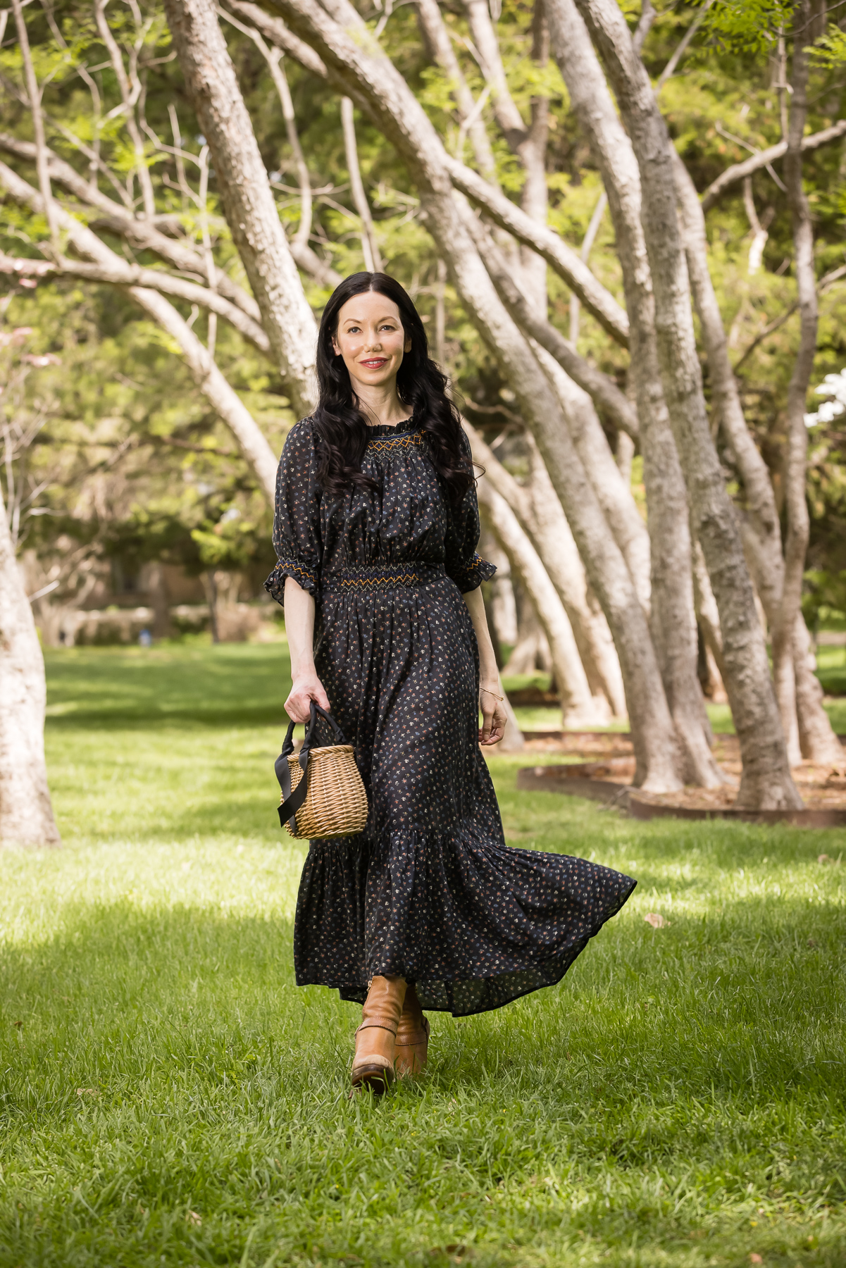 Black Floral Doen Prairie Dress | Prairie Dress by popular Dallas fashion blog, Pretty Little Shoppers: image of a woman walking outside on some grass under some trees and wearing a black Down prairie dress, tan boots, and holding a woven handbag.