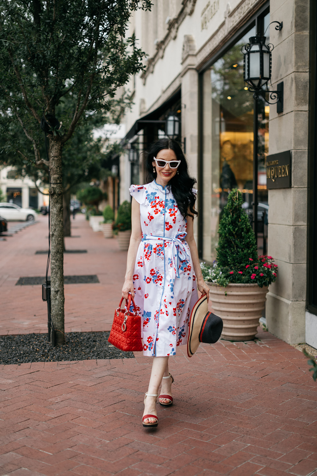 Brunette woman wears Brooks Brothers Shirt Dress with Tommy Hilfiger Sunhat while walking through Highland Park Village in Dallas, Texas. | Brooks Brothers Shirt Dress by popular Dallas fashion blog, Pretty Little Shoppers: image of a woman walking in  Highland Park Village and wearing a red, whites and blue Brooks Brothers shirt dress, white frame sunglasses, and holding a red and black stripe straw hat. 
