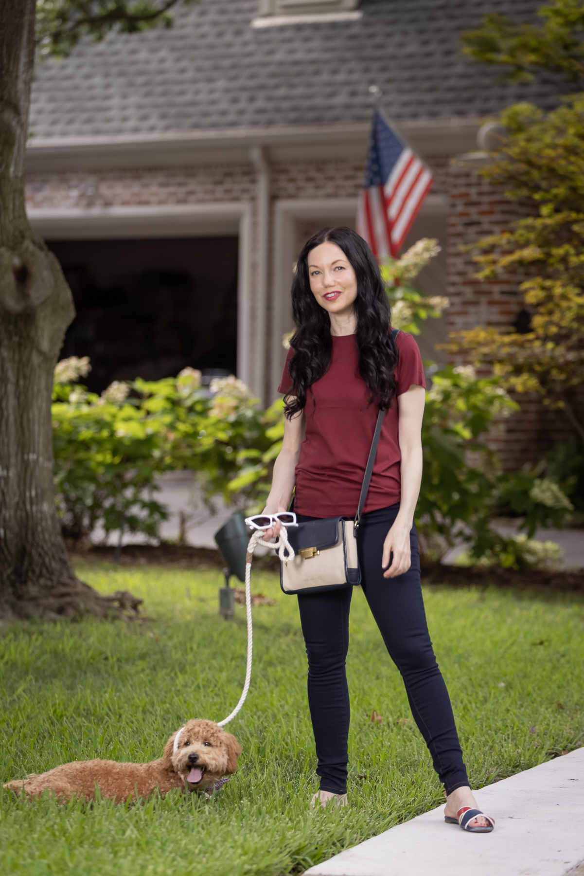 Mott & Bow Jeans and Tee Shirt, Tommy Hilfiger Crossbody Bag, J. Crew Sandals, Italic Cateye Sunglasses, Mini Goldendoodle puppy, Foggy Dog Bowtie, Preppy Style, Dallas Fashion Blogger | Mott and Bow Jeans by popular Dallas fashion blog, Pretty Little Shoppers: image of a woman wearing a crimson t-shirt, Mott and Bow Jeans, blue and red strap slide sandals, and Black and Tan crossbody bag while holding her dog on a leash. 