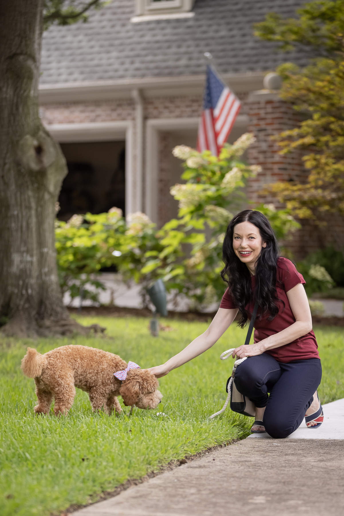 Mott & Bow Jeans and Tee Shirt, Tommy Hilfiger Crossbody Bag, J. Crew Sandals, Italic Cateye Sunglasses, Mini Goldendoodle puppy, Foggy Dog Bowtie, Preppy Style, Dallas Fashion Blogger | Mott and Bow Jeans by popular Dallas fashion blog, Pretty Little Shoppers: image of a woman wearing a crimson t-shirt, Mott and Bow Jeans, blue and red strap slide sandals, and black and tan crossbody bag while kneeling on a sidewalk and petting her doodle breed dog. 