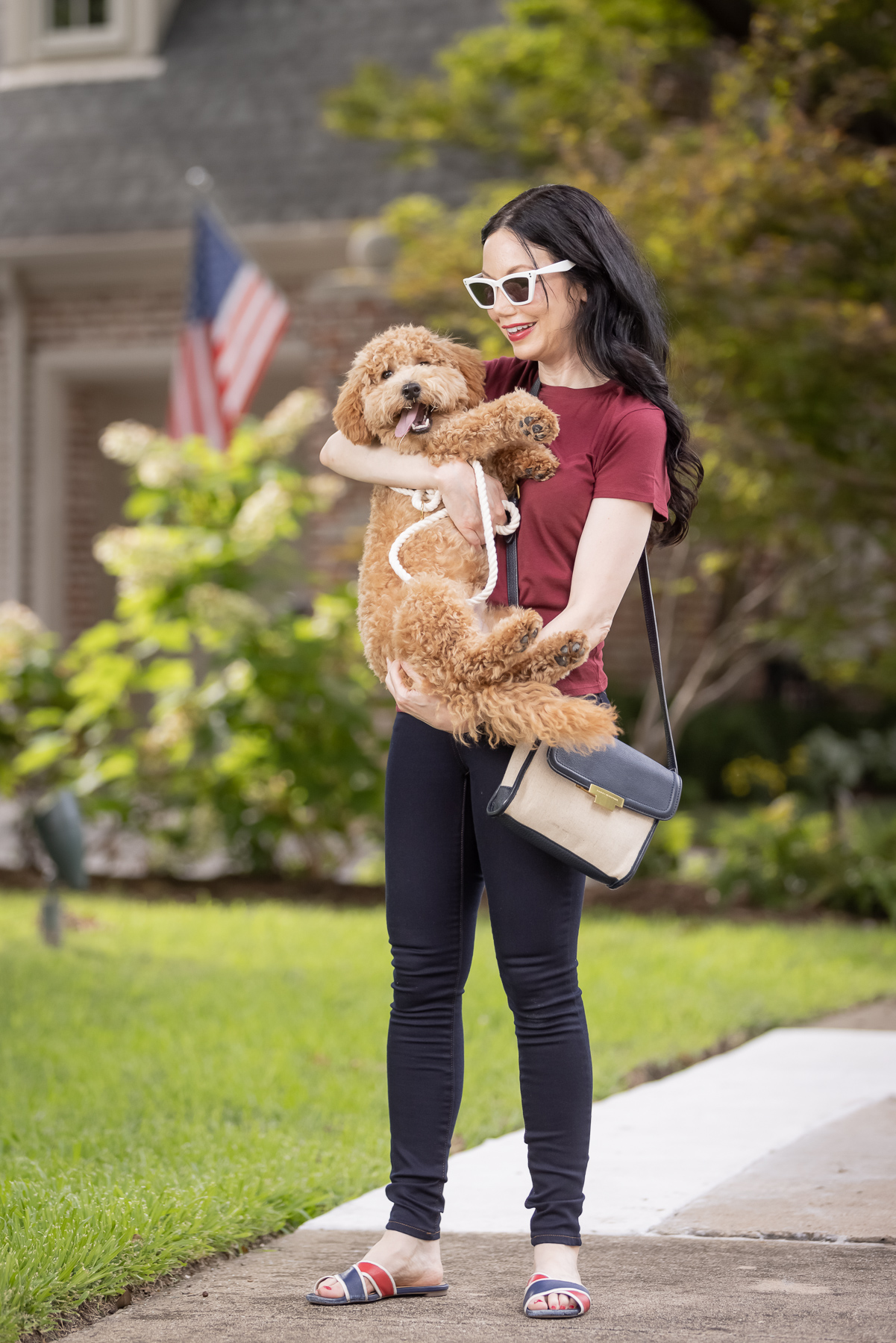 Mott & Bow Jeans and Tee Shirt, Tommy Hilfiger Crossbody Bag, J. Crew Sandals, Italic Cateye Sunglasses, Mini Goldendoodle puppy, Foggy Dog Bowtie, Preppy Style, Dallas Fashion Blogger | Mott and Bow Jeans by popular Dallas fashion blog, Pretty Little Shoppers: image of a woman wearing a crimson t-shirt, Mott and Bow Jeans, blue and red strap slide sandals, and black and tan crossbody bag while standing outside and holding her doodle breed dog. 