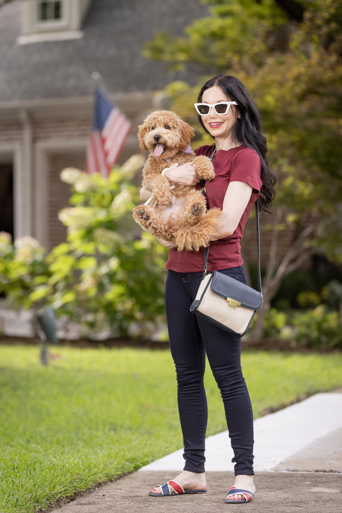 Mott & Bow Jeans and Tee Shirt, Tommy Hilfiger Crossbody Bag, J. Crew Sandals, Italic Cateye Sunglasses, Mini Goldendoodle puppy, Foggy Dog Bowtie, Preppy Style, Dallas Fashion Blogger | Mott and Bow Jeans by popular Dallas fashion blog, Pretty Little Shoppers: image of a woman wearing a crimson t-shirt, Mott and Bow Jeans, blue and red strap slide sandals, and black and tan crossbody bag while holding her doodle breed dog.