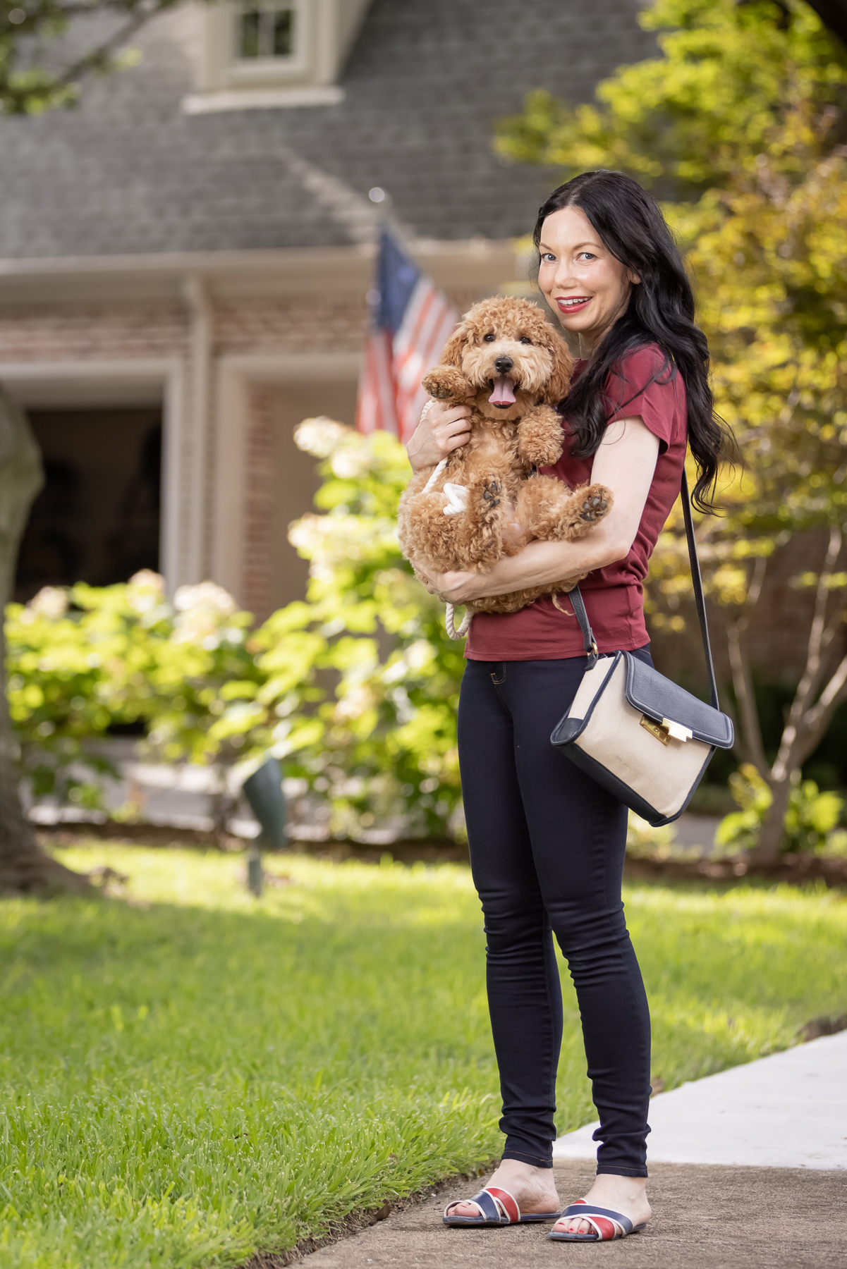 Mott & Bow Jeans and Tee Shirt, Tommy Hilfiger Crossbody Bag, J. Crew Sandals, Italic Cateye Sunglasses, Mini Goldendoodle puppy, Foggy Dog Bowtie, Preppy Style, Dallas Fashion Blogger | Mott and Bow Jeans by popular Dallas fashion blog, Pretty Little Shoppers: image of a woman wearing a crimson t-shirt, Mott and Bow Jeans, blue and red strap slide sandals, and black and tan crossbody bag while standing outside and holding her doodle breed dog. 