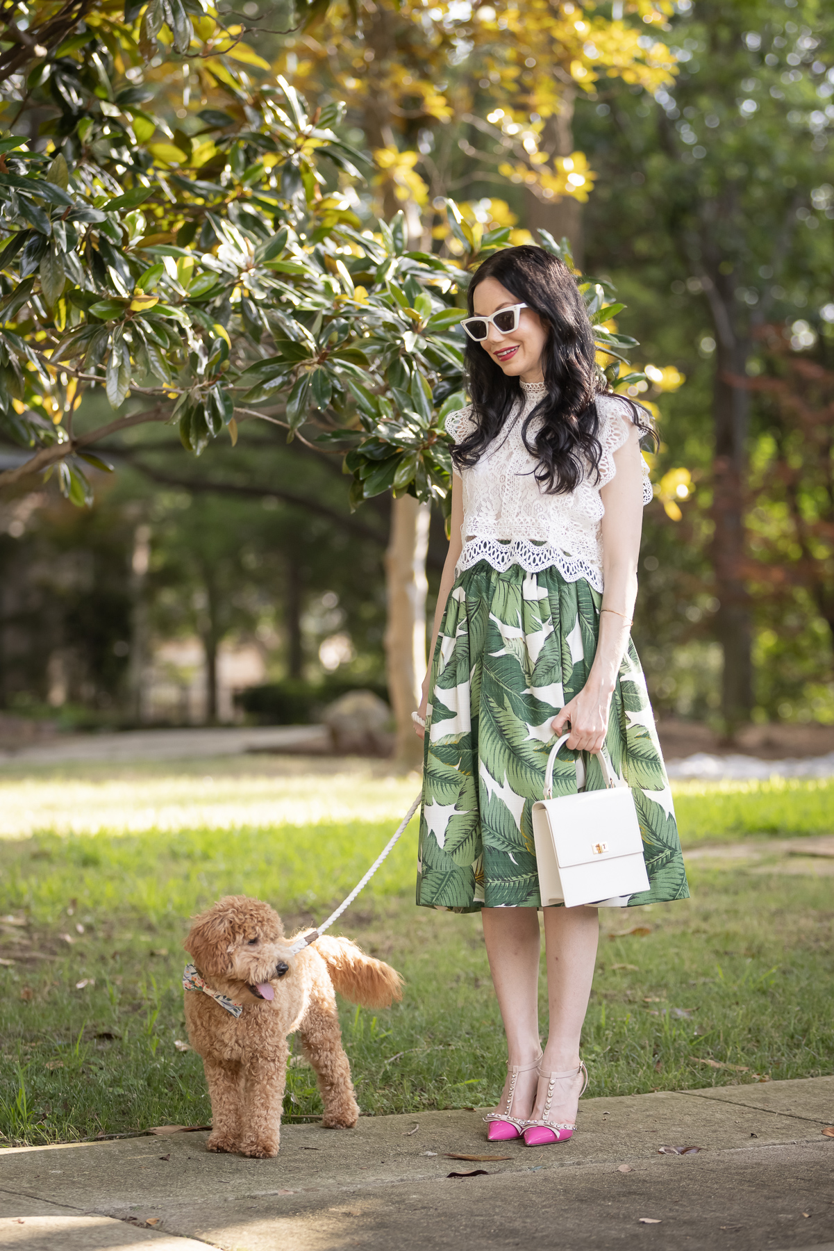 Palm Print Skirt, Neely & Chloe Bag, White Lace Top, Pink Pumps, Fashion Blogger, Fall Transitional Style, Mini Goldendoodle puppy, Fashion Blogger | Palm Print Skirt by popular Dallas fashion blog, Pretty Little Shoppers: image of a woman walking her dog outside and wearing a palm print skirt, lace cropped top, and pink heels. 