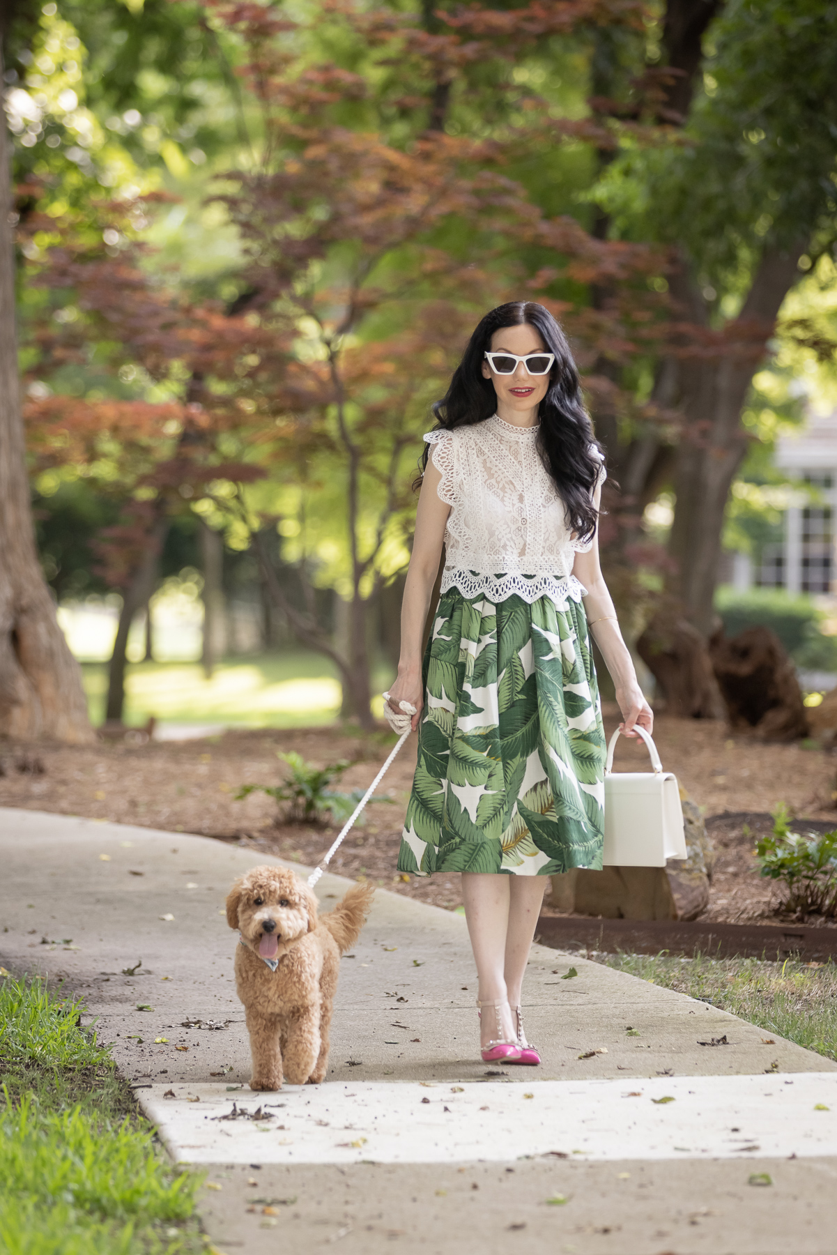 Palm Print Skirt, Neely & Chloe Bag, White Lace Top, Pink Pumps, Fashion Blogger, Fall Transitional Style, Mini Goldendoodle puppy, Fashion Blogger, | Palm Print Skirt by popular Dallas fashion blog, Pretty Little Shoppers: image of a woman walking her dog outside and wearing a palm print skirt, lace cropped top, and pink heels. 