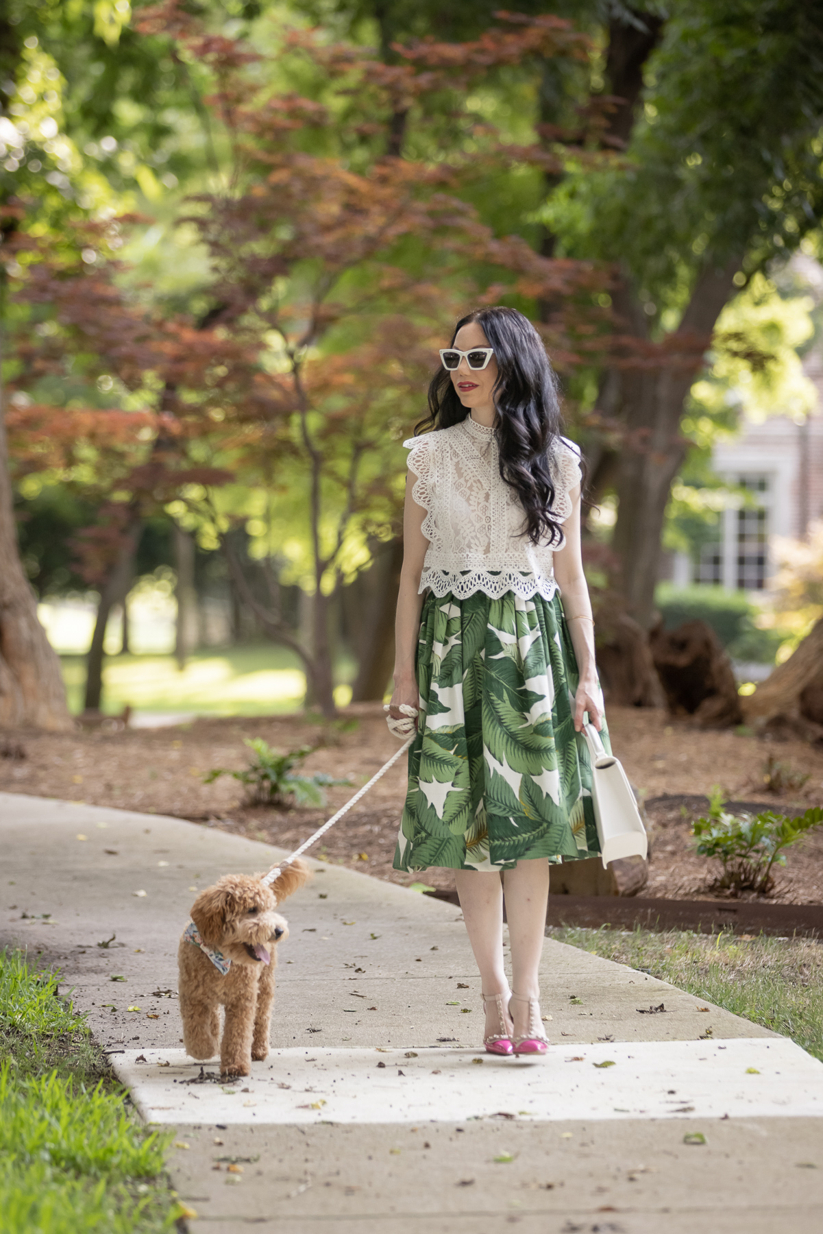 Palm Print Skirt, Neely & Chloe Bag, White Lace Top, Pink Pumps, Fashion Blogger, Fall Transitional Style, Mini Goldendoodle puppy, Fashion Blogger | Palm Print Skirt by popular Dallas fashion blog, Pretty Little Shoppers: image of a woman walking her dog outside and wearing a palm print skirt, lace cropped top, and pink heels. 