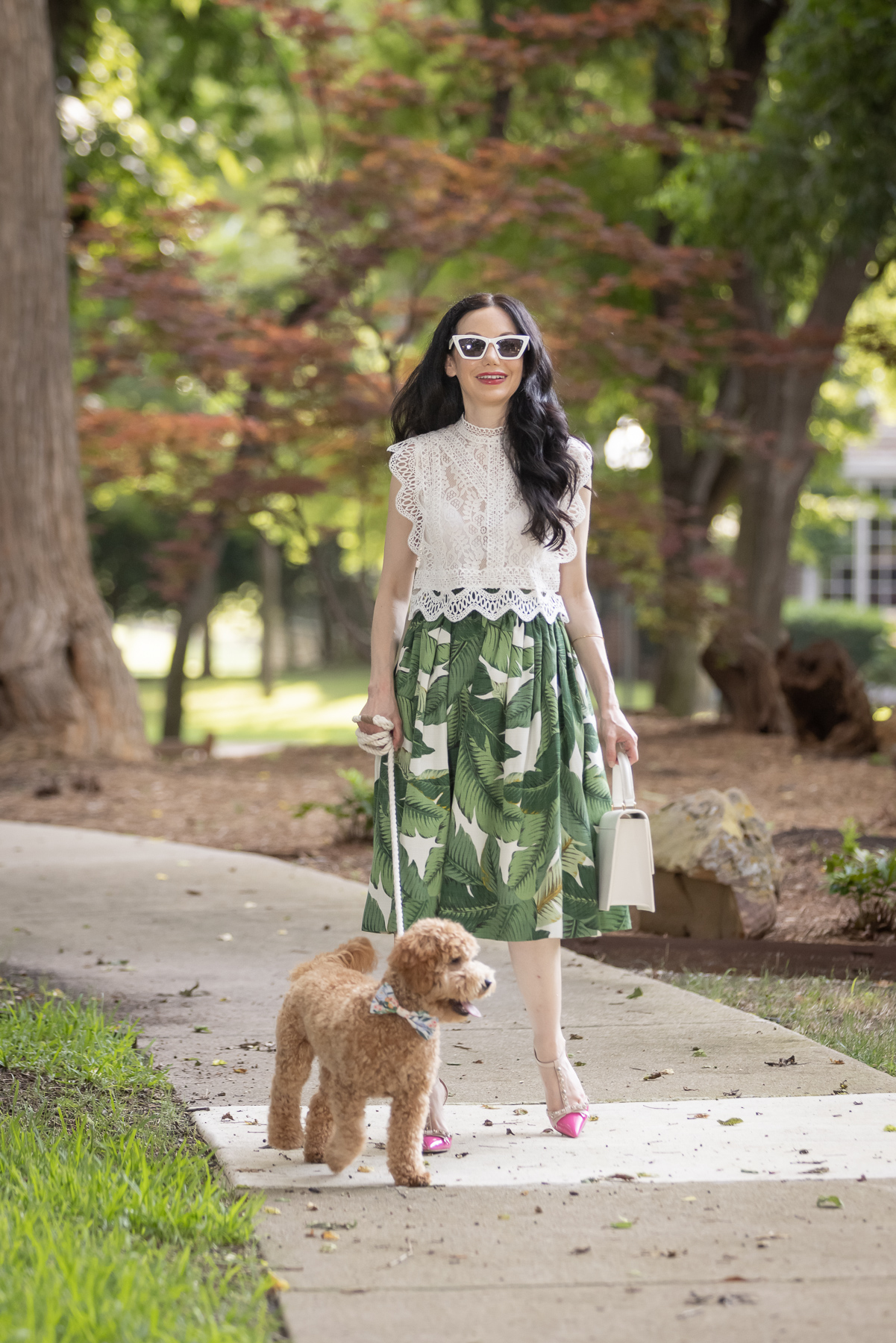 Palm Print Skirt, Neely & Chloe Bag, White Lace Top, Pink Pumps, Fashion Blogger, Fall Transitional Style, Mini Goldendoodle puppy, Fashion Blogger | Palm Print Skirt by popular Dallas fashion blog, Pretty Little Shoppers: image of a woman walking her dog outside and wearing a palm print skirt, lace cropped top, and pink heels. 