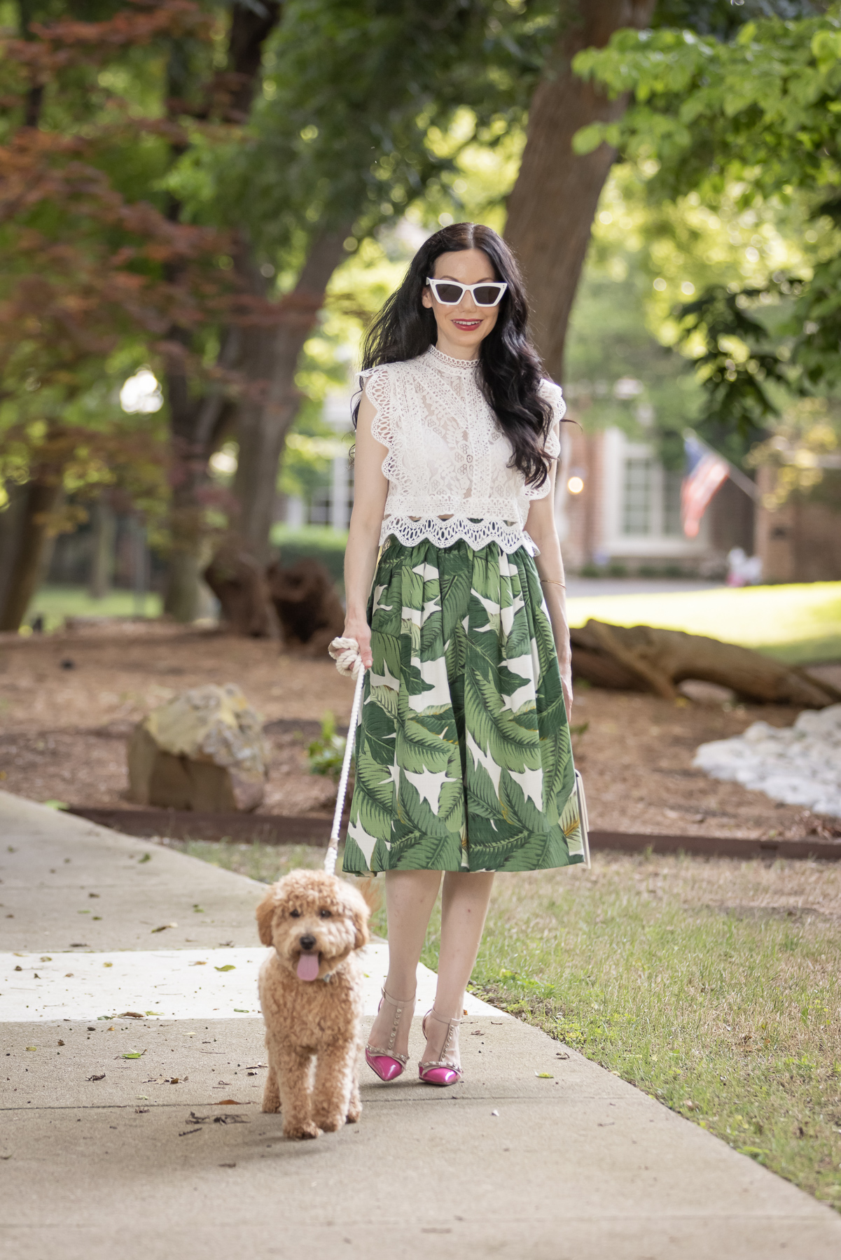 Palm Print Skirt, Neely & Chloe Bag, White Lace Top, Pink Pumps, Fashion Blogger, Fall Transitional Style, Mini Goldendoodle puppy, Fashion Blogger | Palm Print Skirt by popular Dallas fashion blog, Pretty Little Shoppers: image of a woman walking her dog outside and wearing a palm print skirt, lace cropped top, and pink heels. 