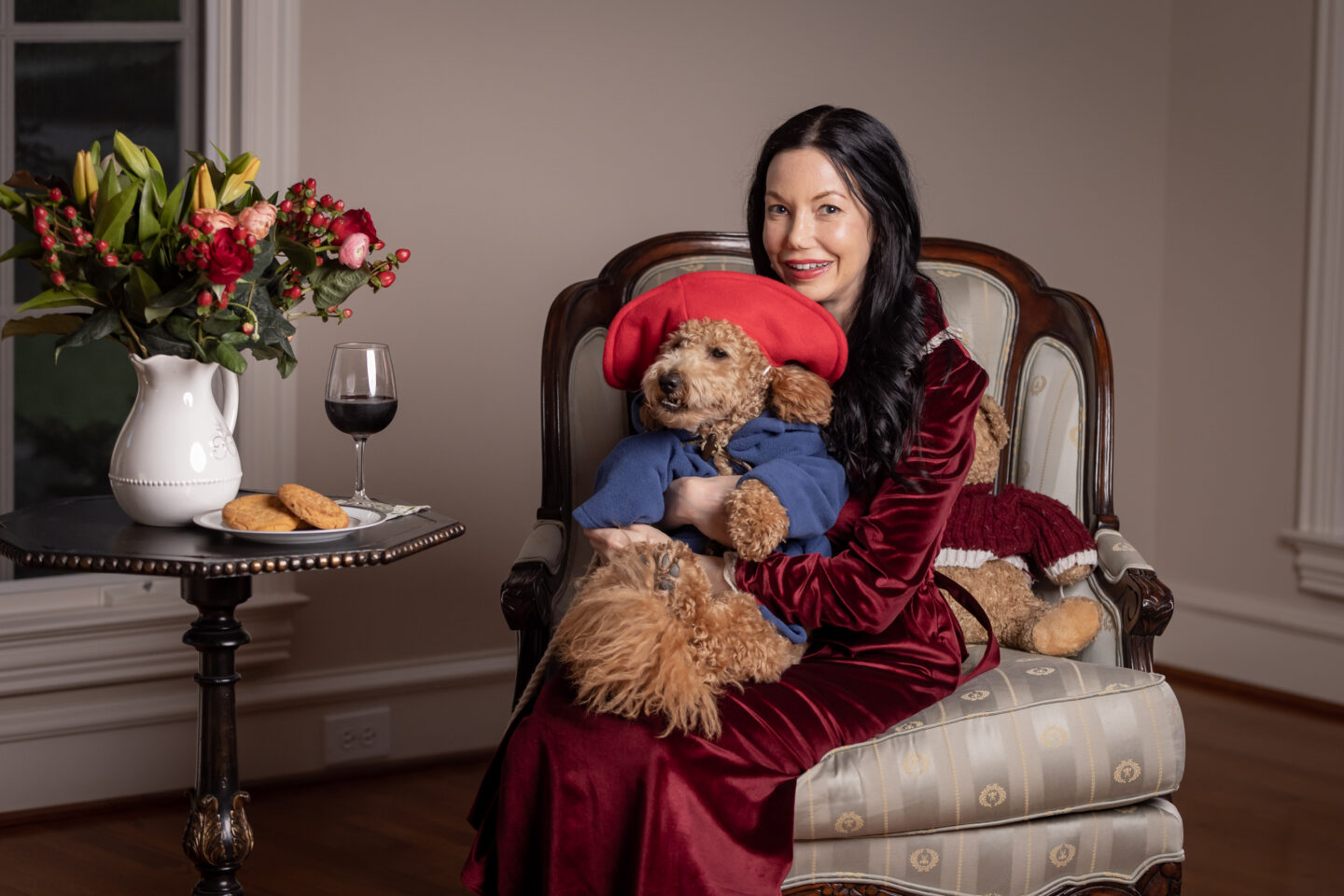Brunette Woman wearing Simple Retro Red Velvet Dress holds her Goldendoodle Puppy dressed as Paddington Bear